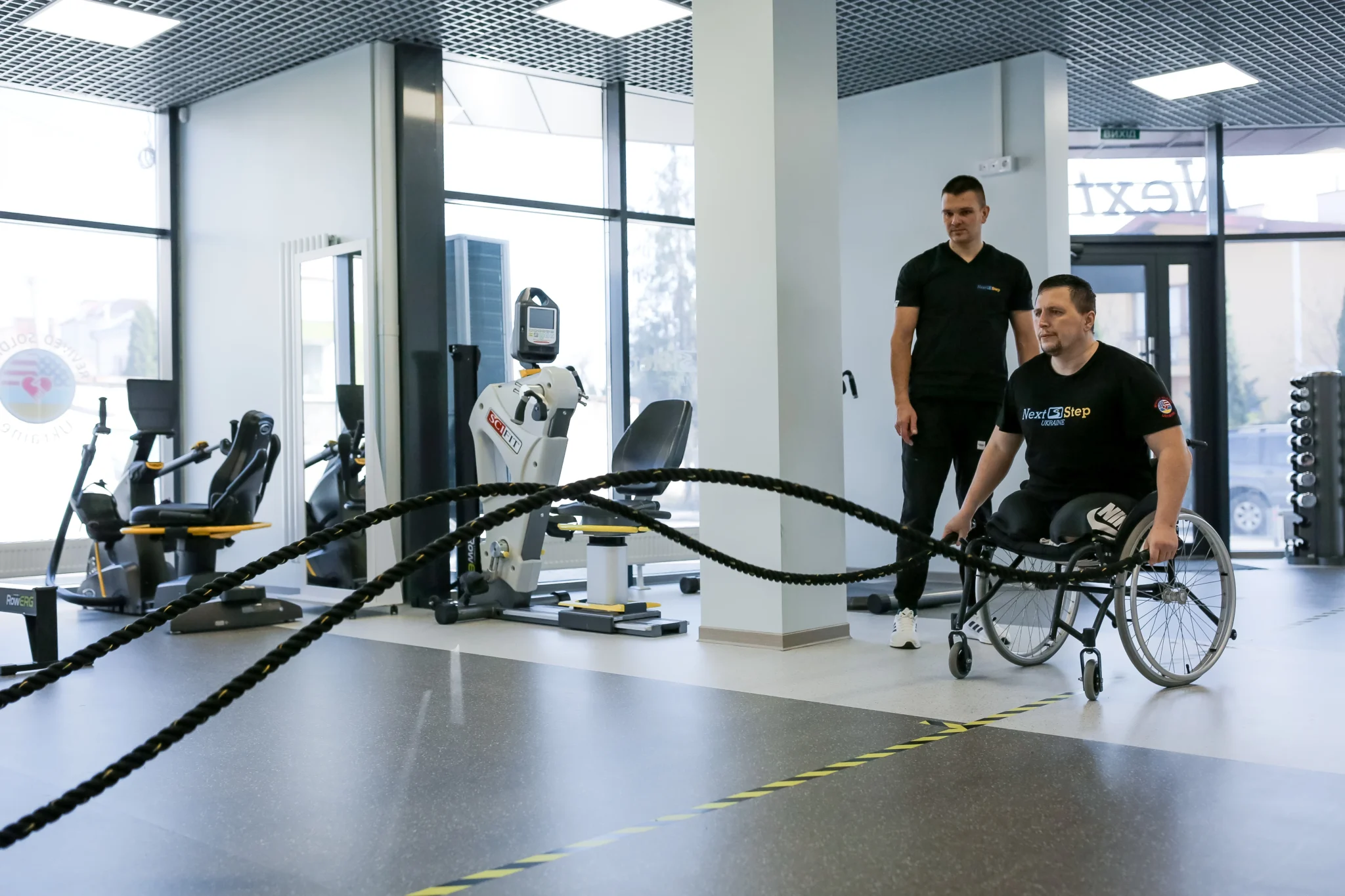 Salle de rééducation : un homme en fauteuil roulant s’exerce à côté d’un entraîneur
